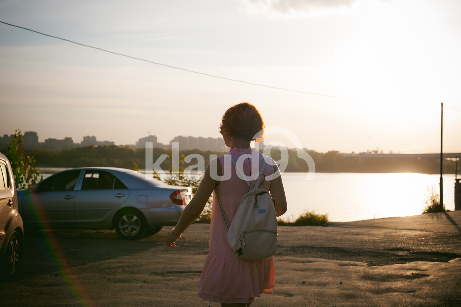 girl in a pale pink dress with red hair and a backpack goes to his car in the parking lot by the river, sunset on a summer day