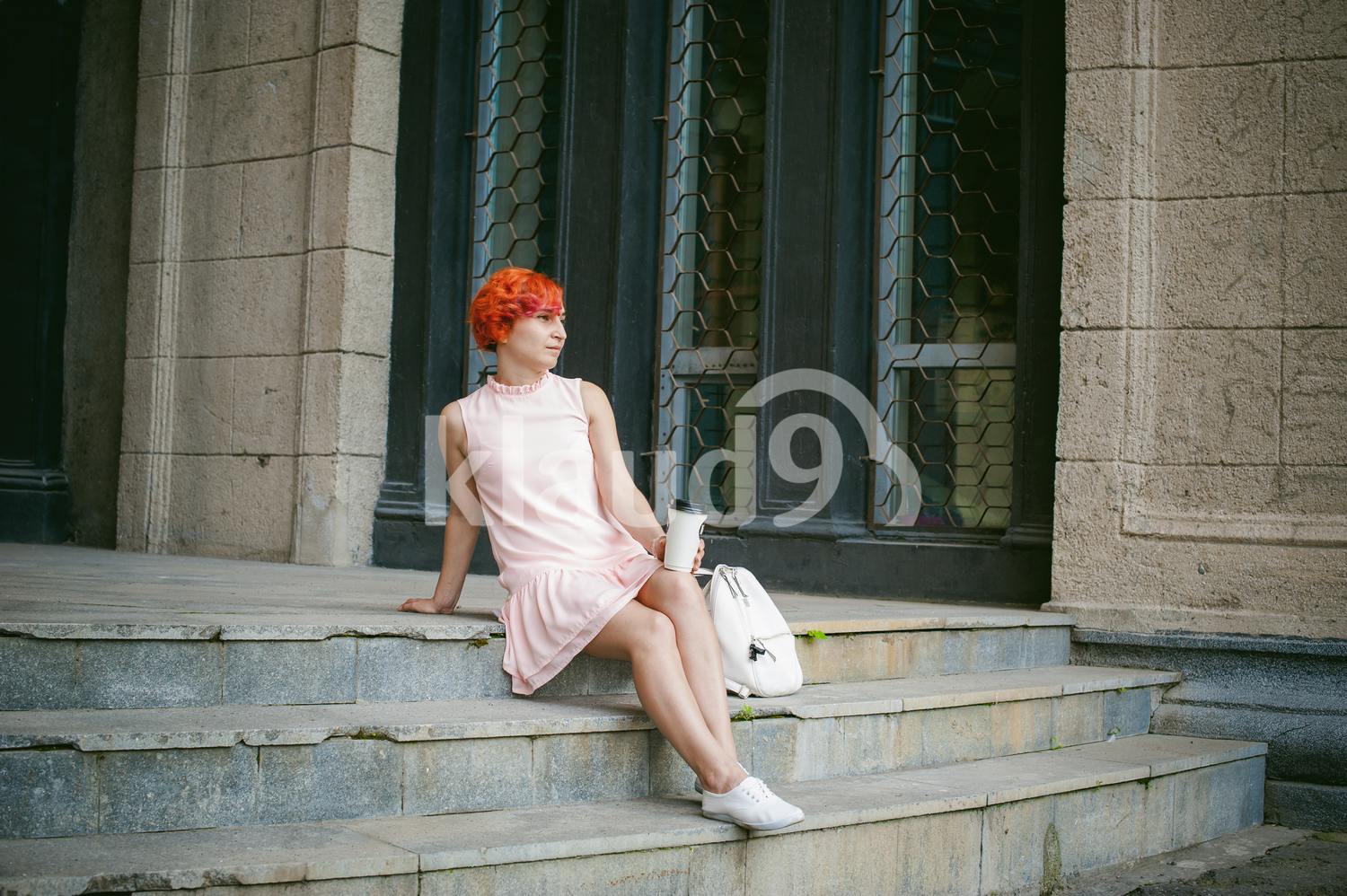 drinks coffee from a cup. woman with dyed red hair in a pale pink dress drinking coffee, holding a paper cup of coffee to go, sitting on the steps outside a summer day.